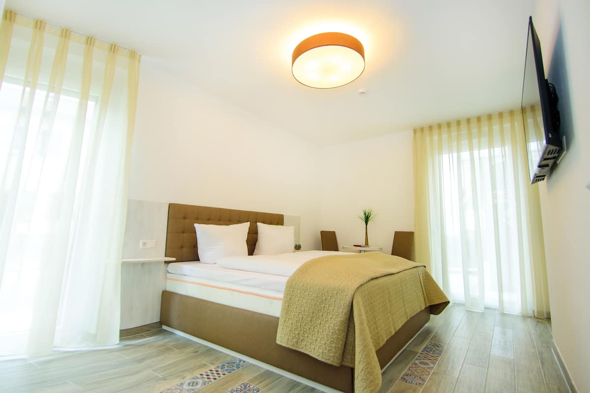 A hotel room with a bed, curtains, a flat-screen TV mounted on the wall, and a ceiling light fixture.