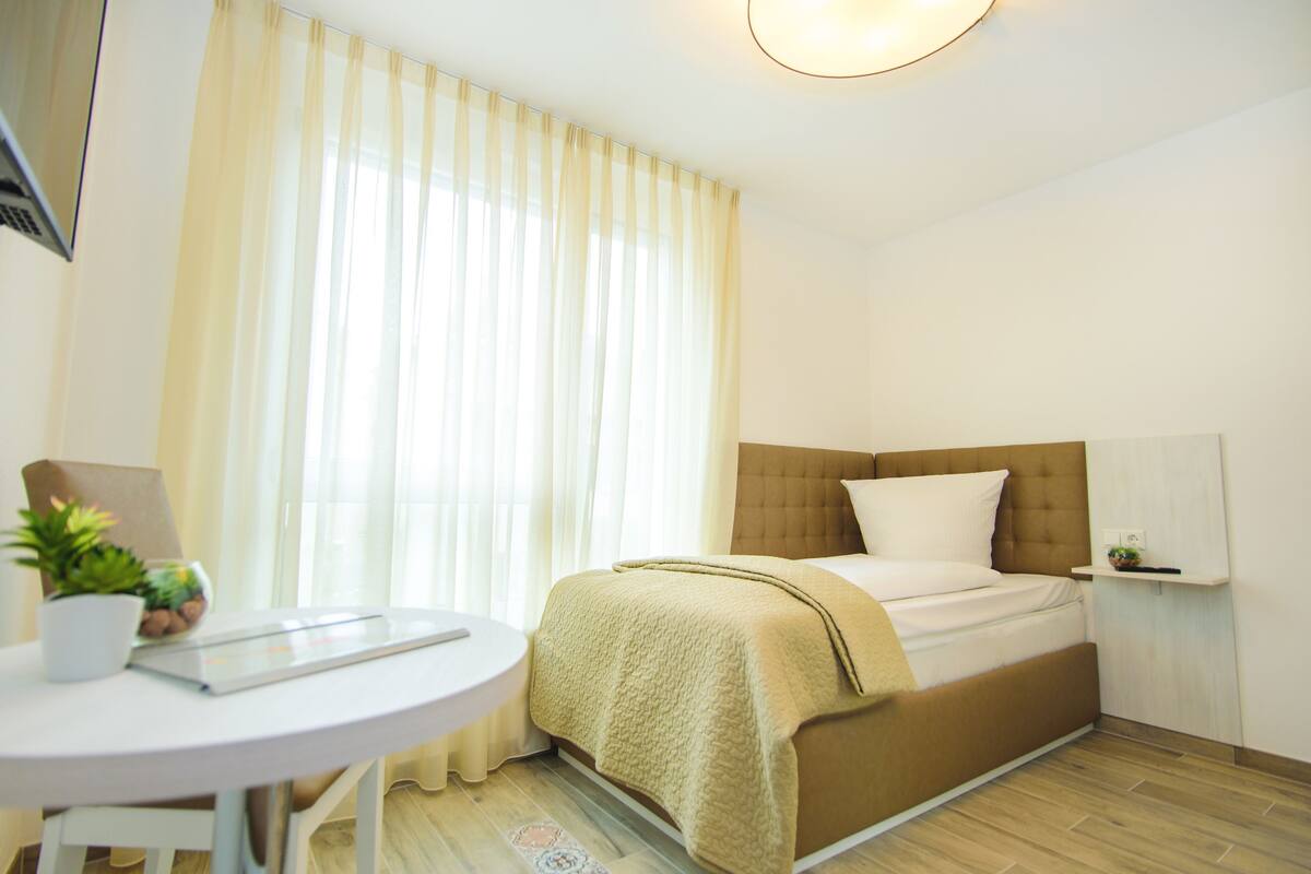 A hotel room with a bed, a round table, a chair, a potted plant, and a window with curtains.