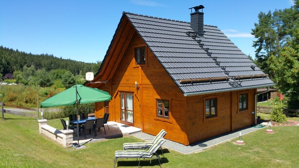 A wooden house with a sloped roof, a patio with a table and chairs, and two lounge chairs. - Where nature lives ... enjoy your vacation. (Drognitz)