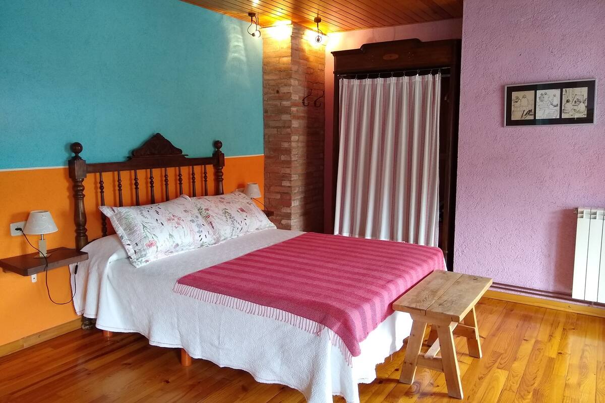 Un dormitorio con cama de madera, una mesita de noche con lámpara, un banco, pared de ladrillo, ventana con cortinas y un cuadro colgado en la pared.