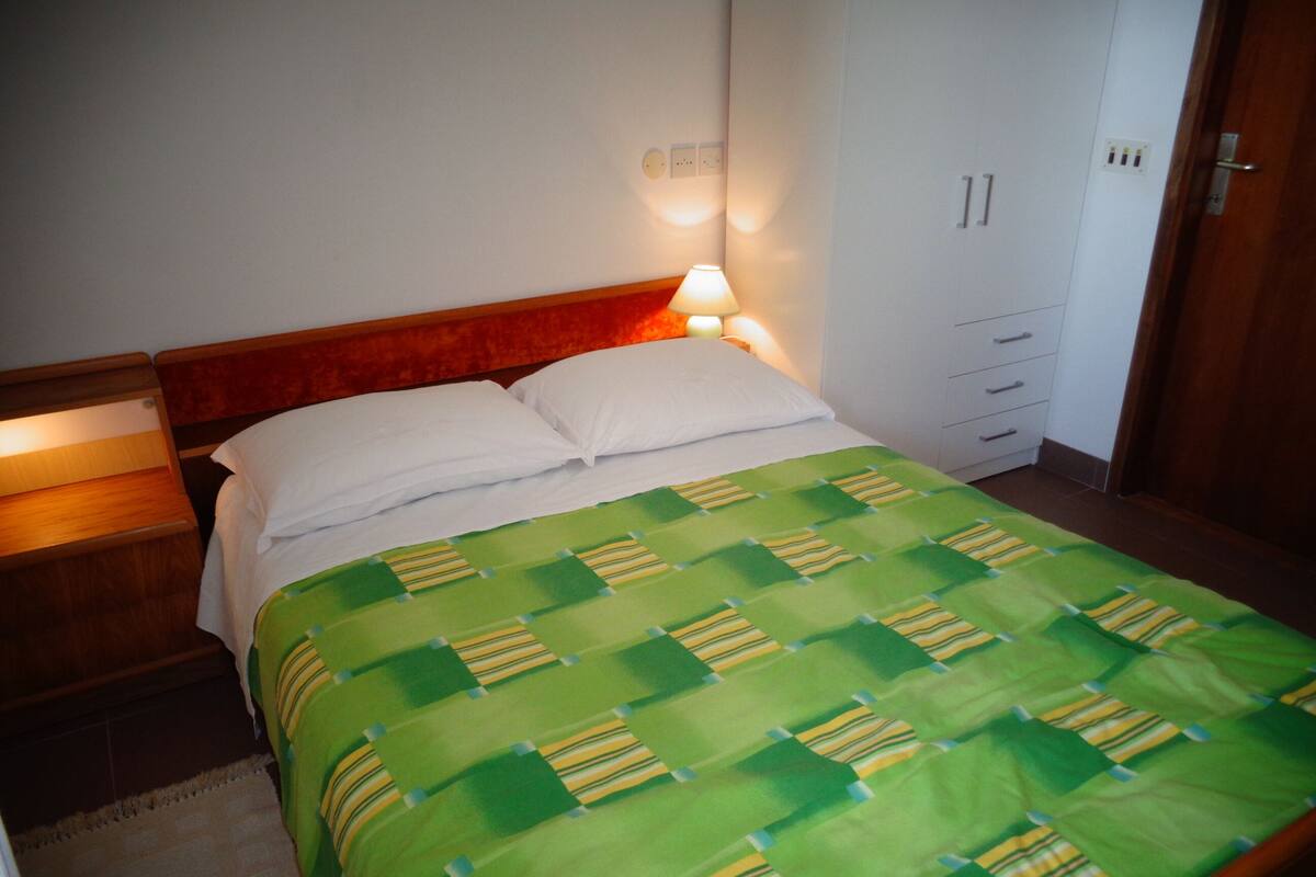 A neatly made bed with a green and yellow striped bedspread, a wooden headboard, and a bedside lamp.