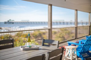 A table with a bottle and glasses, blue chairs, and a view of the ocean and pier. - 211 E Arctic Ave - Tee "Fore" Two - Oceanfront Beach House with Private Beach Access! (Folly Beach)