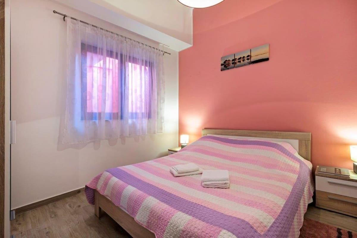 Une chambre à coucher avec un lit, une fenêtre ornée de rideaux légers, une lampe de chevet et un tableau accroché au mur.