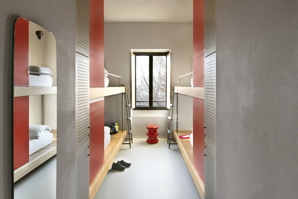 Un couloir étroit, avec des lits superposés de chaque côté, une fenêtre au fond et un tabouret rouge au centre.