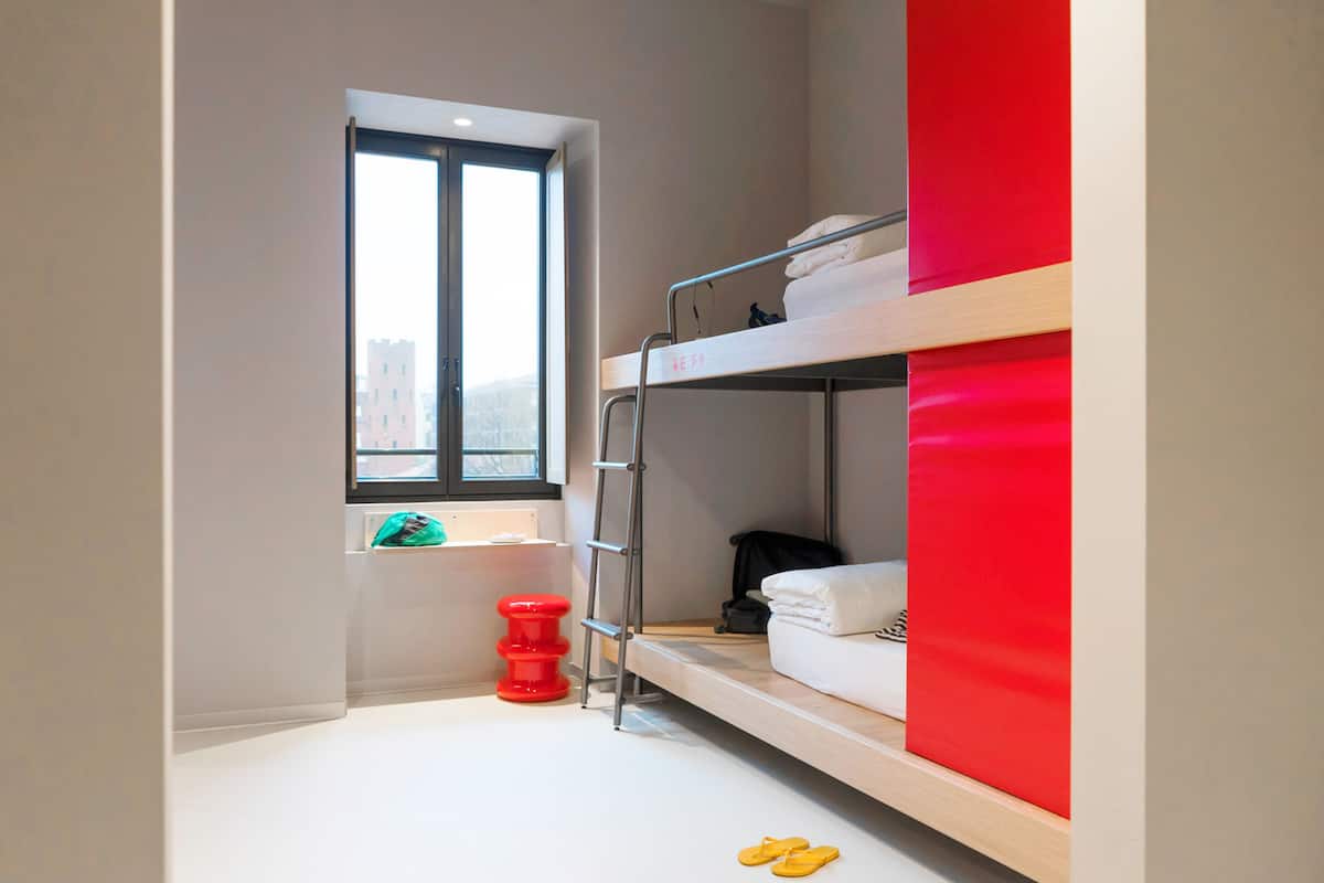 Une chambre avec un lit superposé, une fenêtre, un tabouret rouge et une paire de pantoufles jaunes posées sur le sol.