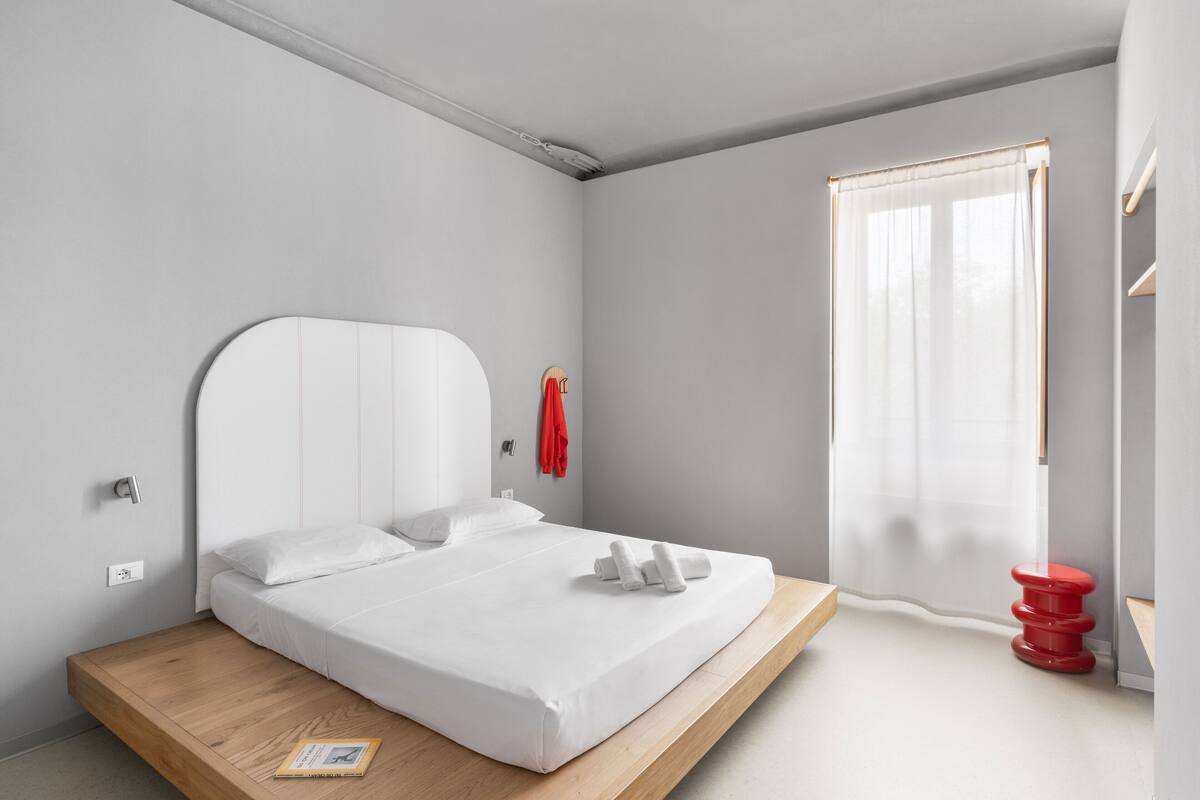 Une chambre à coucher moderne avec un lit surélevé en bois, du linge de lit blanc, un tabouret rouge et une fenêtre agrémentée de rideaux légers.