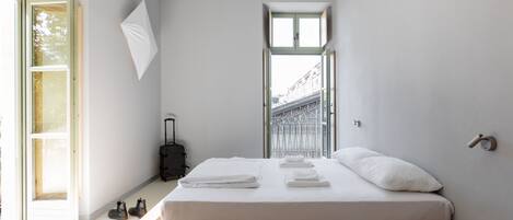 A minimalist hotel room with a bed, a wooden desk, a laptop, and a suitcase.
