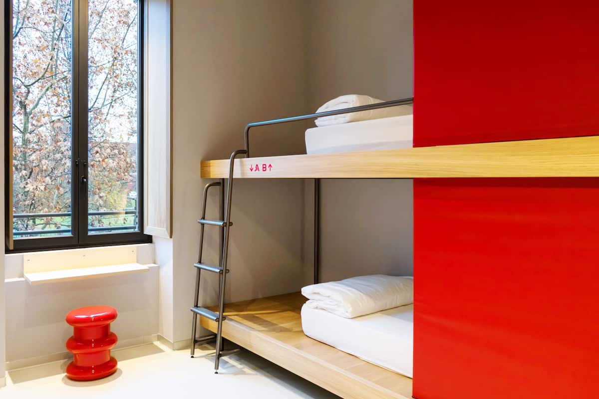 Un lit superposé avec une échelle, une fenêtre donnant sur les arbres et un tabouret rouge.