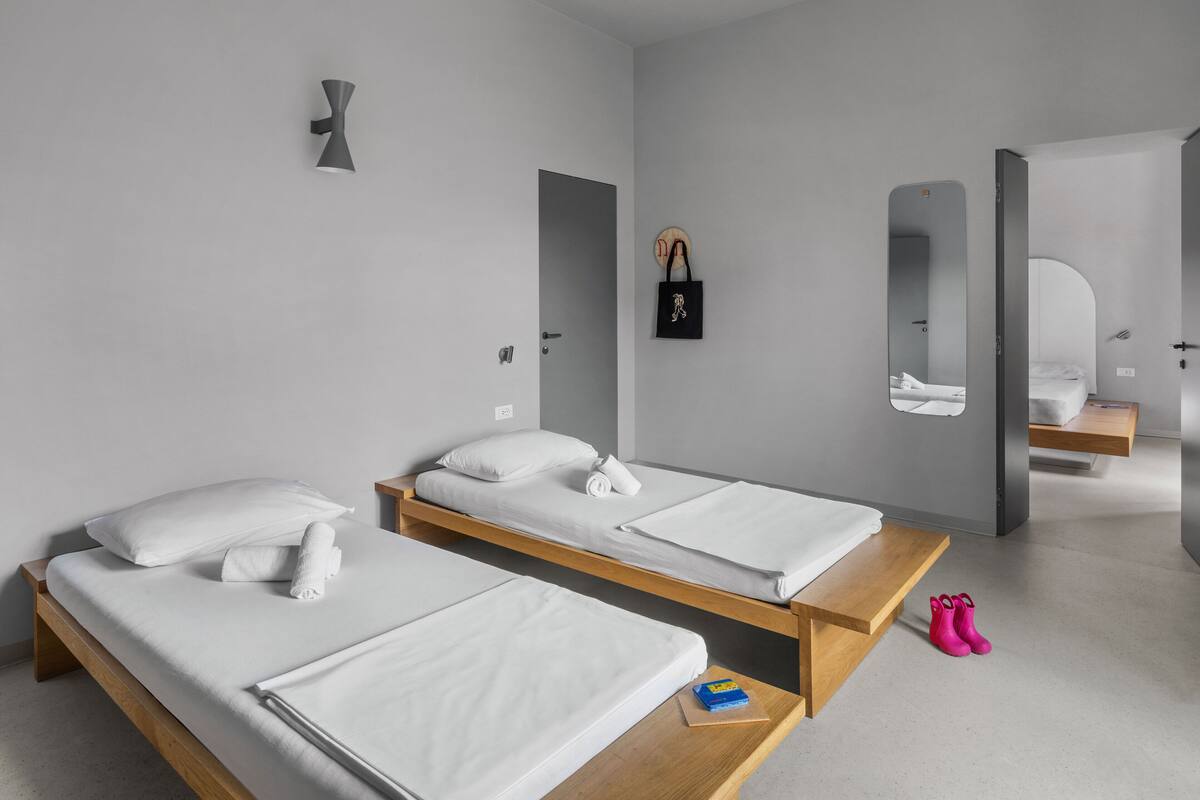Une chambre d’hôtel moderne équipée de deux lits simples, d’un banc en bois, d’une petite table, d’un miroir et d’un porte-bagages fixé au mur.