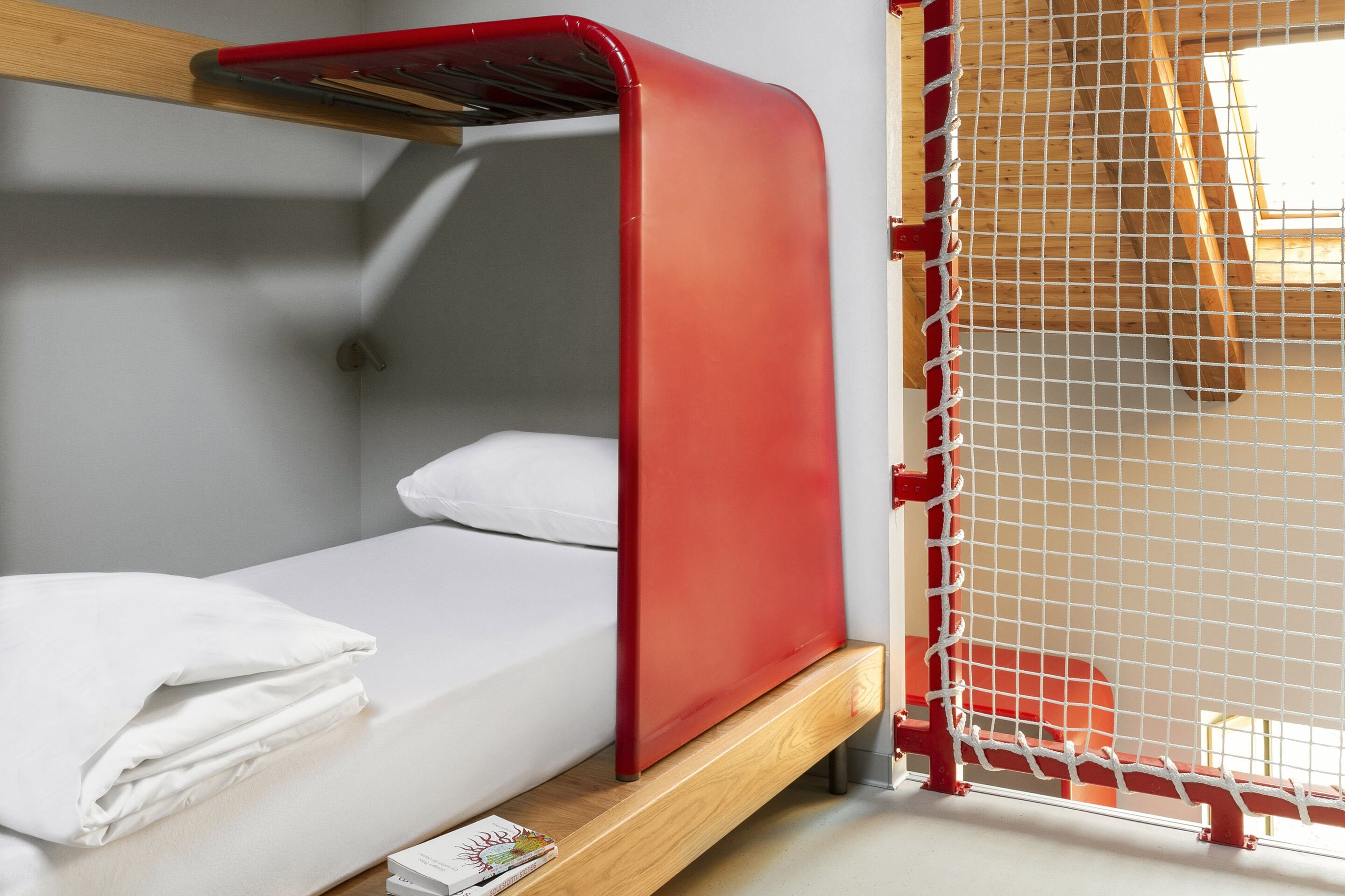 Una cama litera con estructura roja y ropa de cama blanca, una mesita con un libro, y una ventana con estructura de madera.