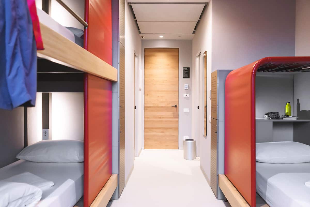 Un couloir bordé de lits superposés de chaque côté, une porte en bois au fond et une poubelle au milieu.
