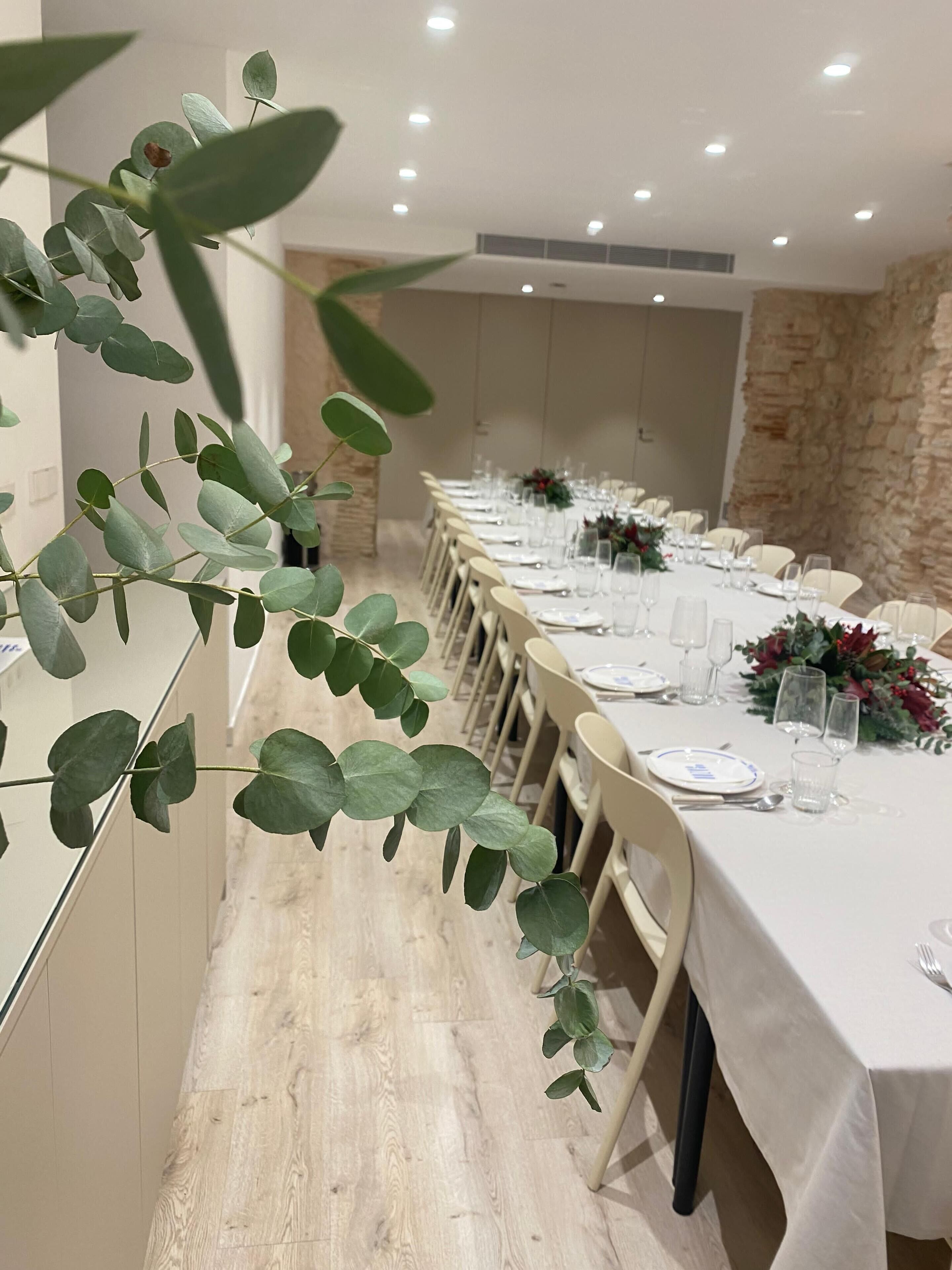 Una larga mesa de comedor dispuesta con manteles blancos, sillas y cristalería, en una habitación con pared de piedra y suelo de madera.