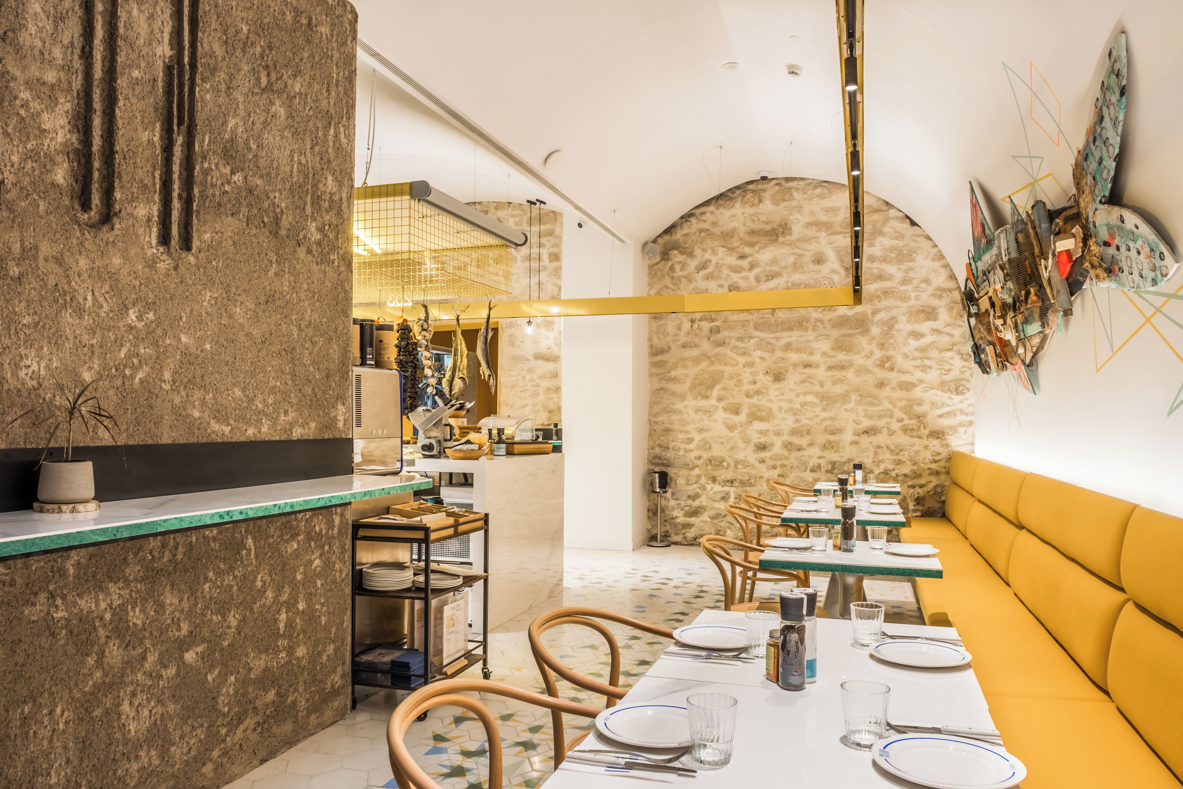 Un restaurante con pared de piedra, sillas de madera y un banco amarillo. Las mesas están dispuestas con platos y vasos.