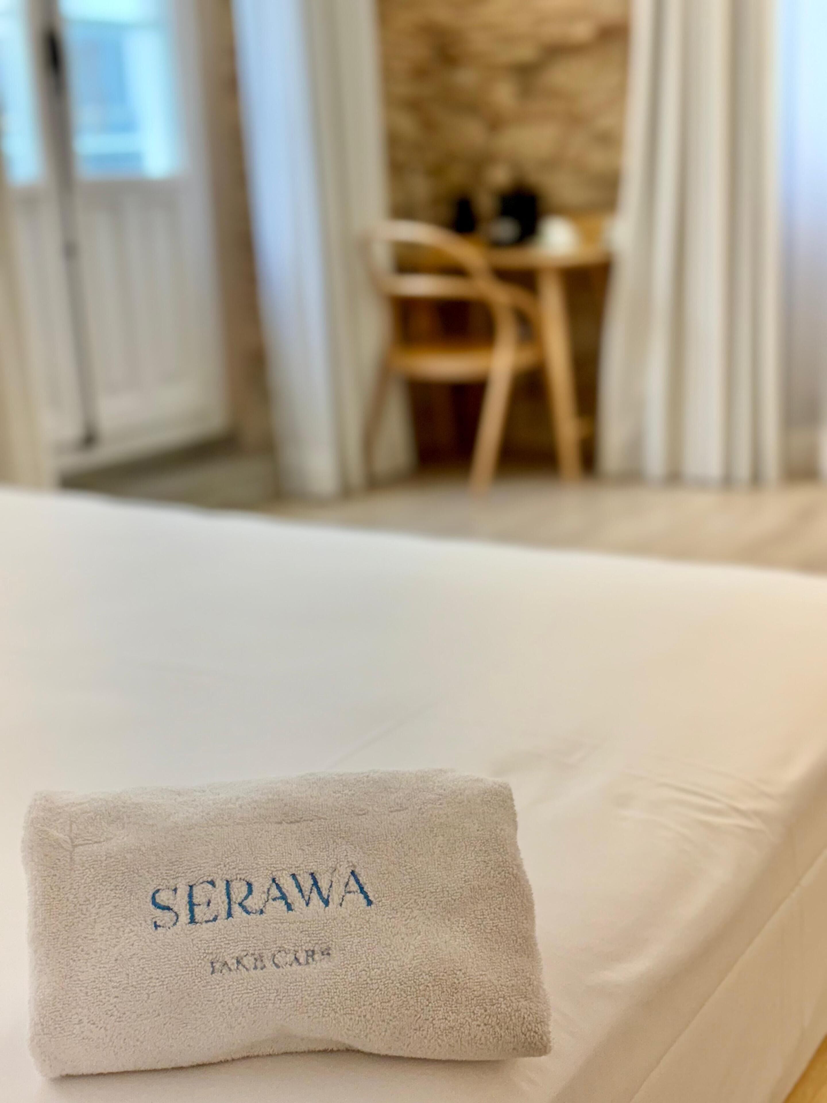 A neatly made bed with a folded towel on it, a wooden chair, and a stone wall in the background.