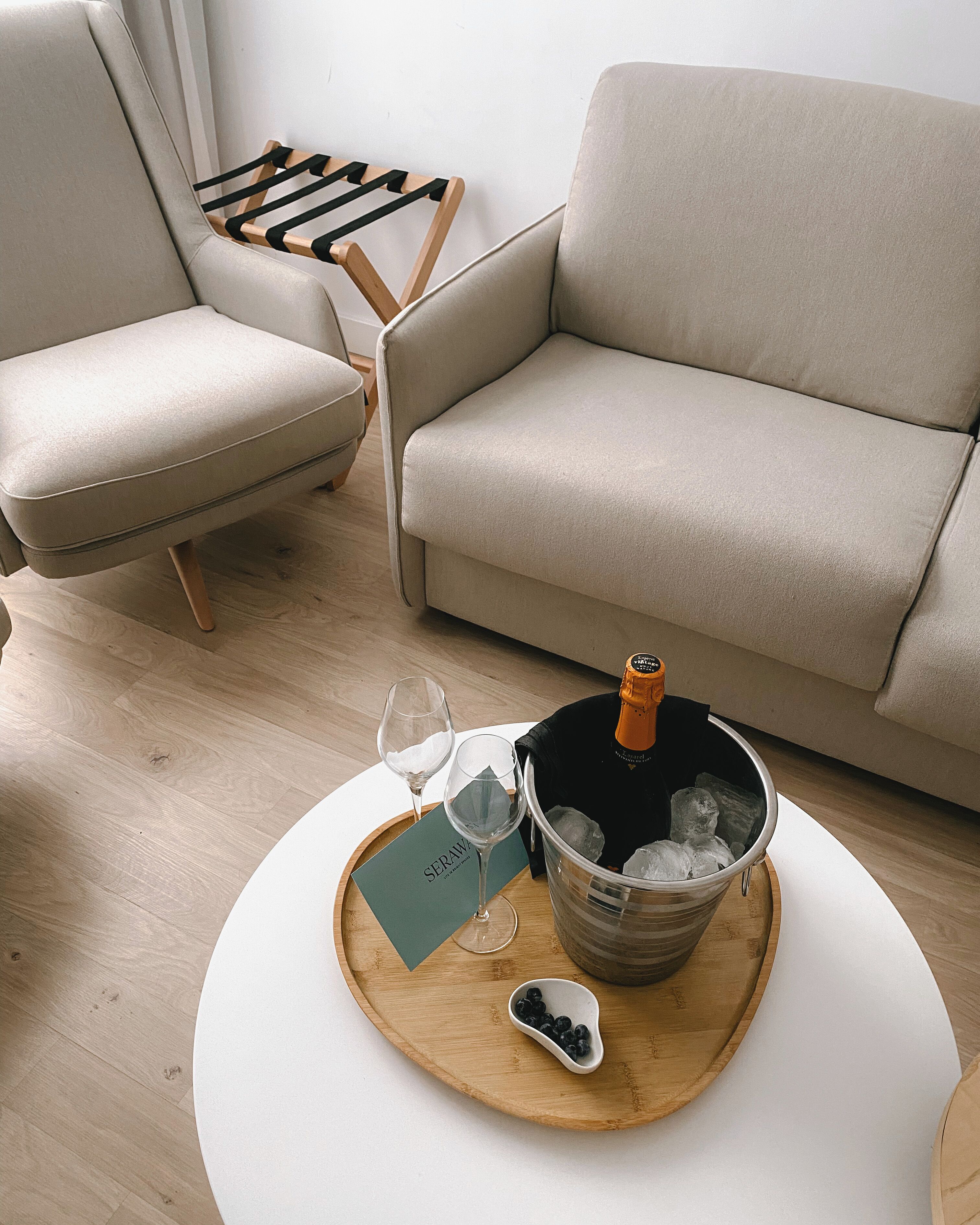 A modern living room with a beige sofa, a wooden coffee table, and a chair. On the table, there is a bottle of champagne, two wine glasses, and a small bowl of blueberries.