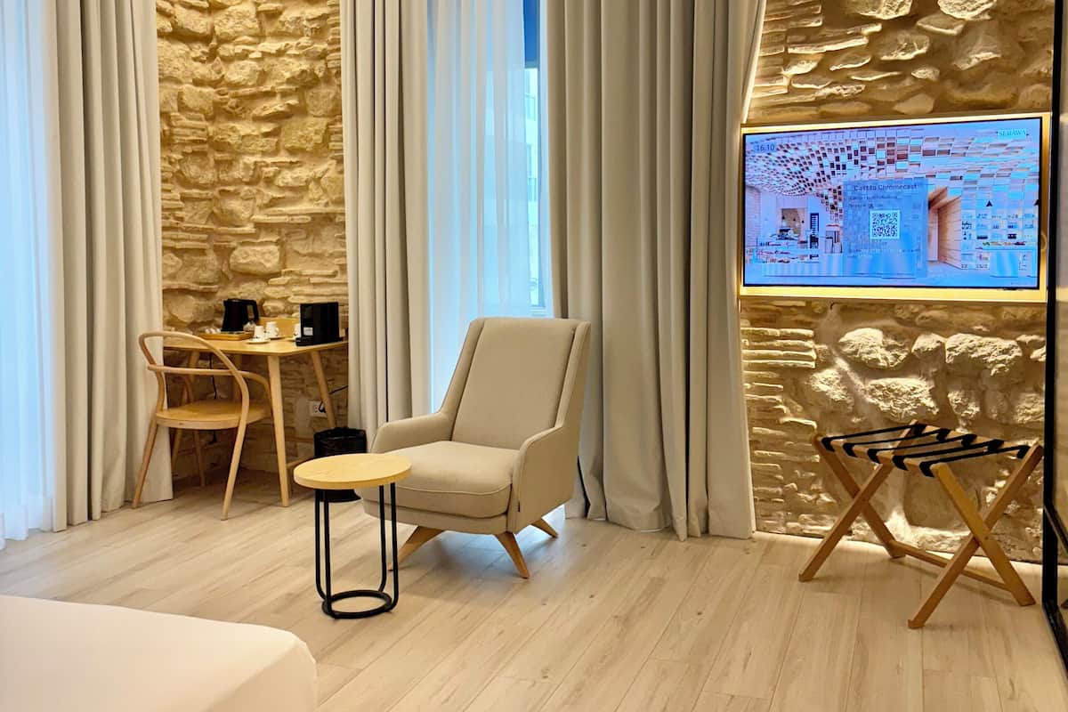 Een moderne hotelkamer met een stenen muur, een bed, een stoel, een tafeltje en een televisiescherm waarop een plattegrond van de kamer wordt weergegeven.