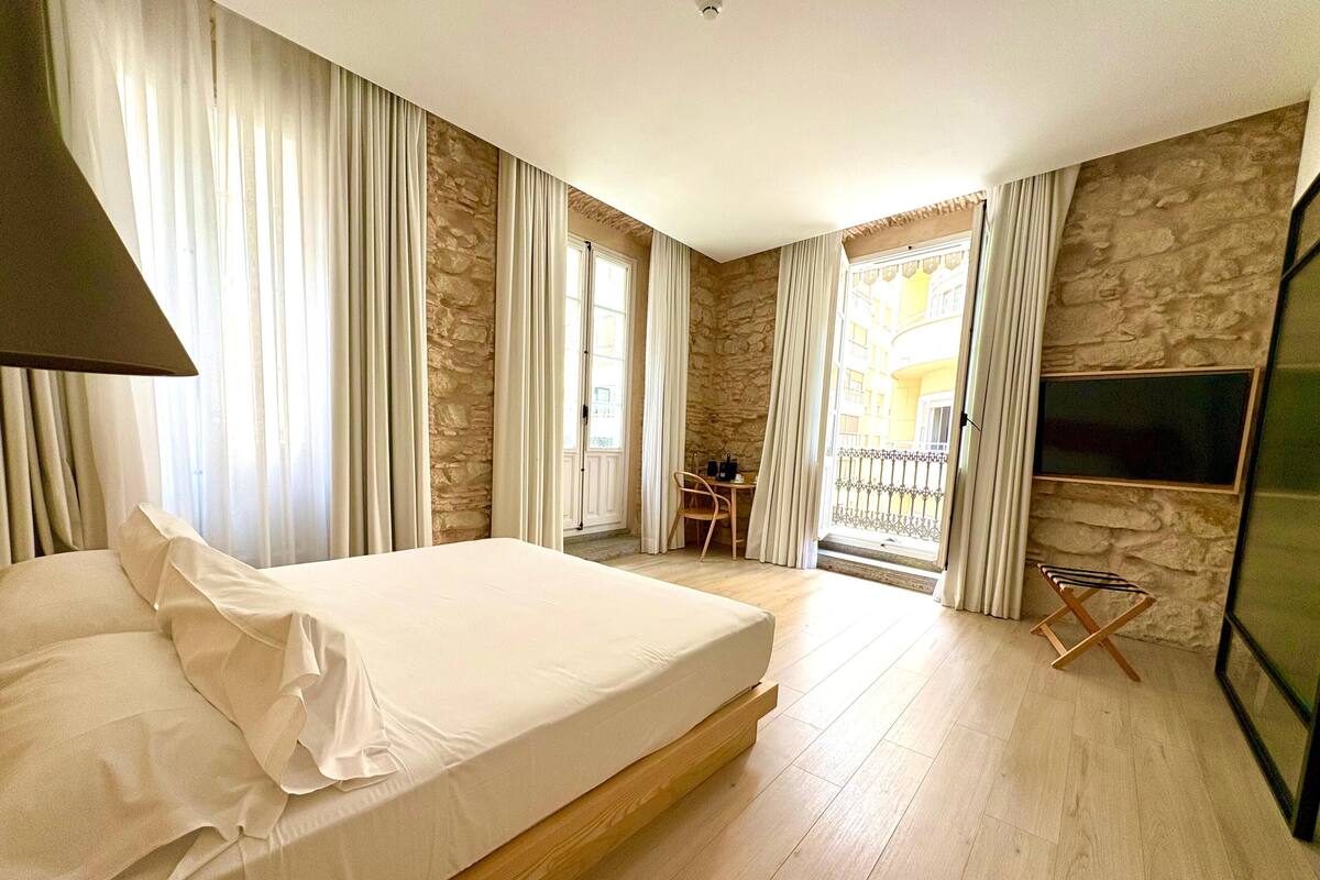 Hotelkamer met een groot bed, stenen muren, een balkon met tafel en stoel, en een aan de muur bevestigde televisie.