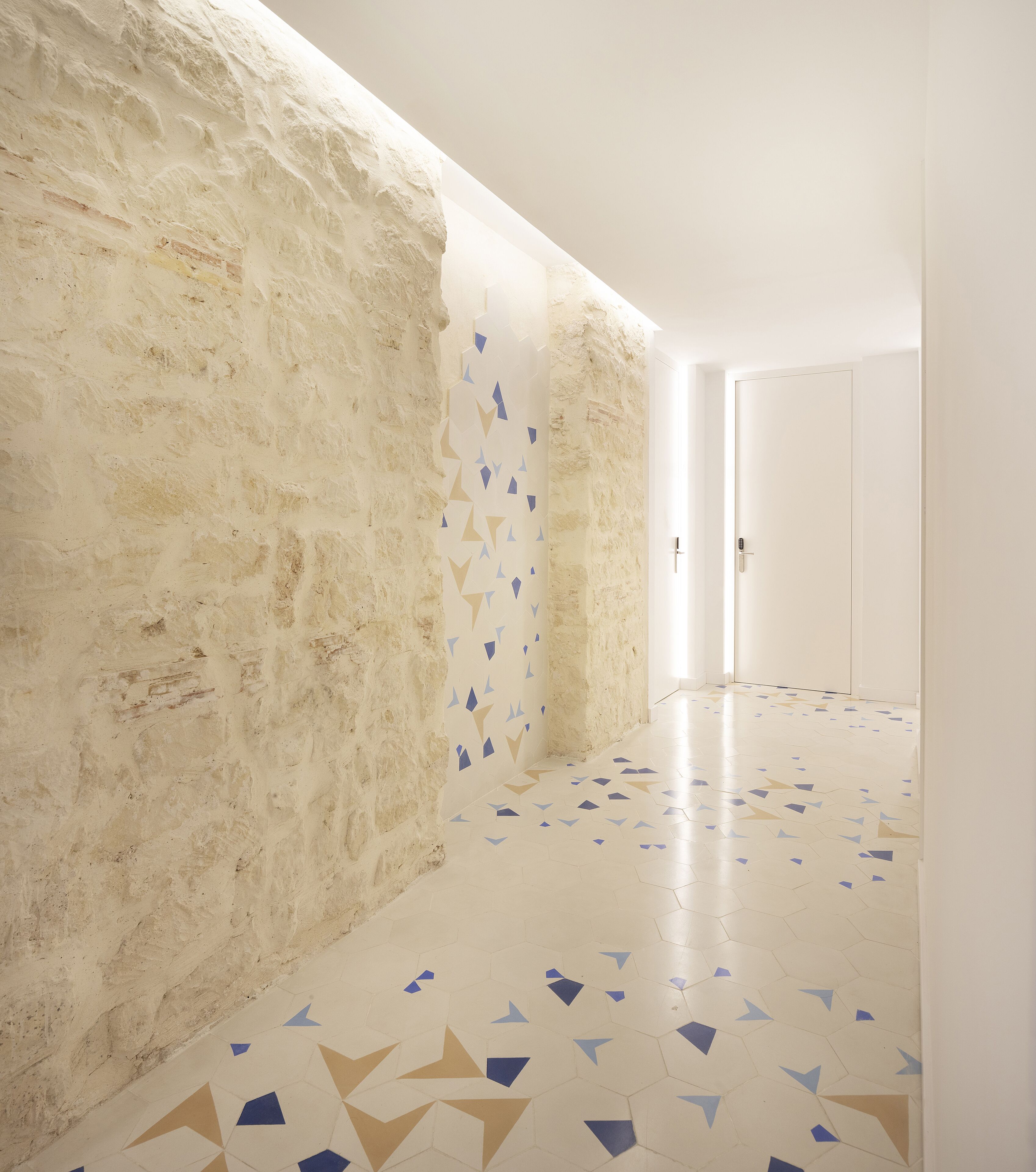 Un couloir avec un mur en pierre à la texture particulière, un sol orné de motifs géométriques et une porte blanche sobre au fond.