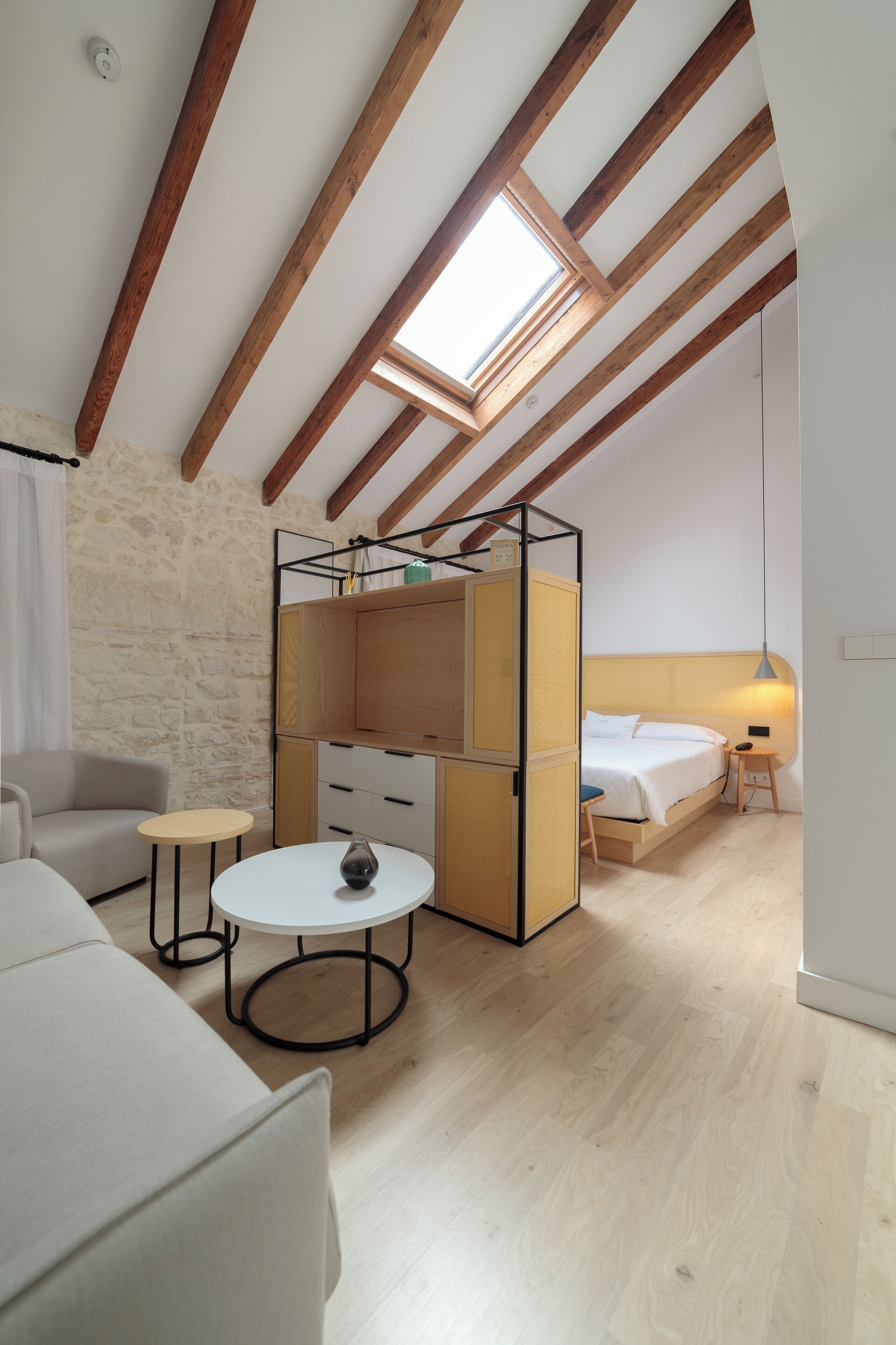 A loft bedroom with exposed wooden beams, a skylight, a bed, a wardrobe, a sofa, and a small round table.