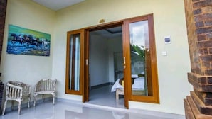 A room with a sliding glass door leading to a balcony, a painting on the wall, and two wicker chairs. - Bali Billy Village (Canggu)