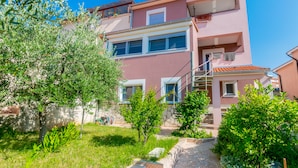 A multi-story building with a pink facade, balconies, and a garden area with trees and shrubs. - Affordable Apartment Bruno in Pula (Pula)