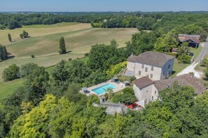A large house with a swimming pool, surrounded by lush greenery and fields.