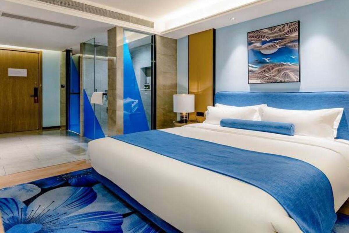 A modern hotel room with a large bed, a nightstand, a wall-mounted painting, and a glass partition.