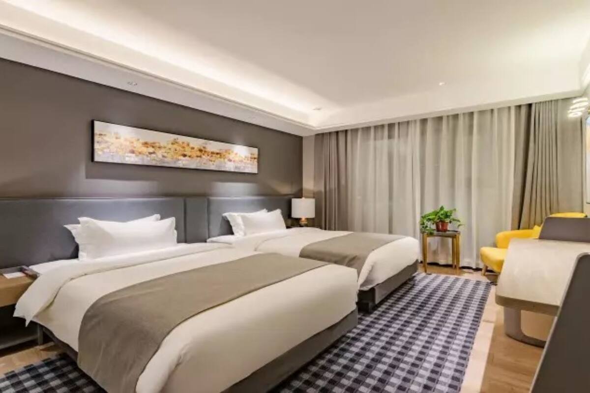 A hotel room with two beds, a large window with curtains, a small table with a plant, and a chair.