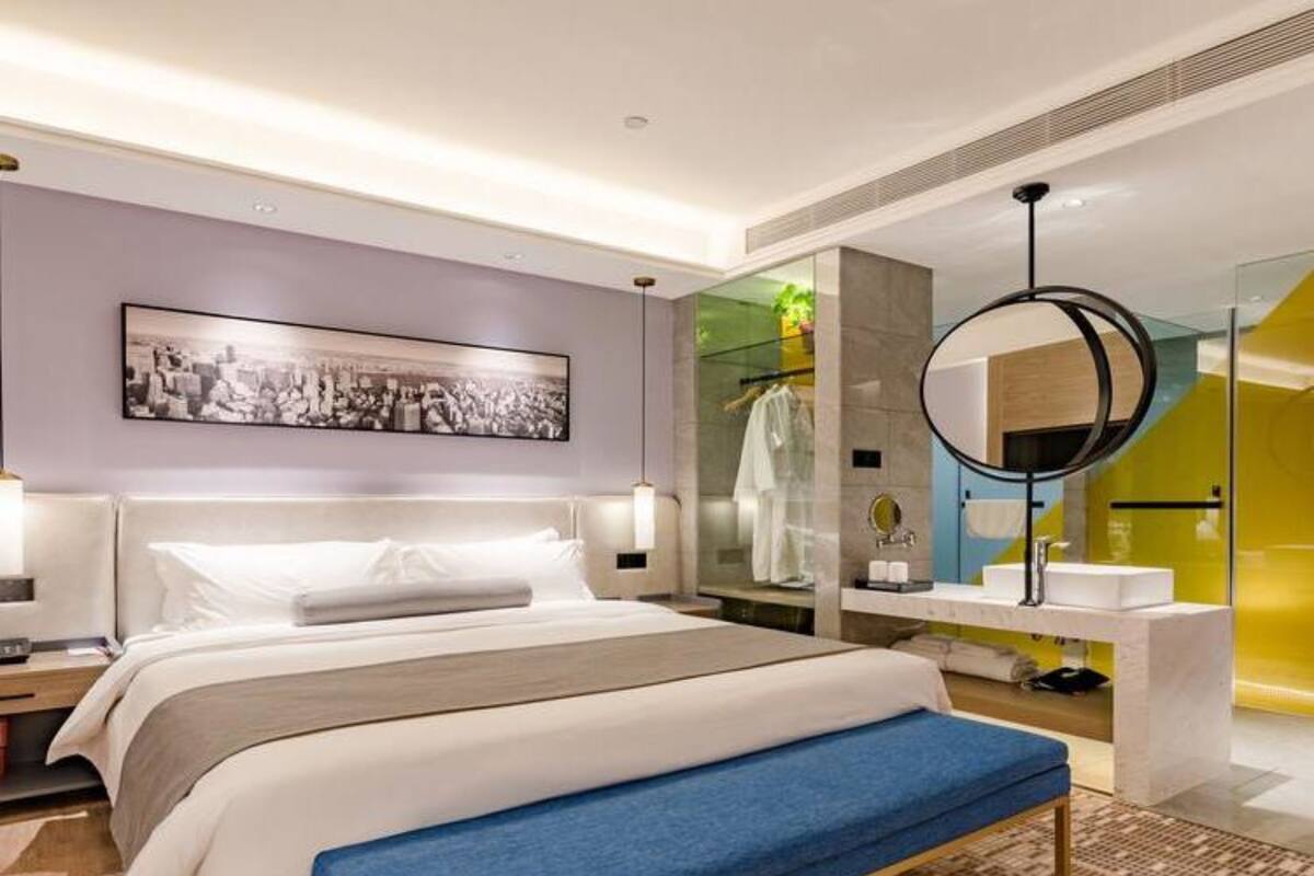 A modern hotel room with a large bed, a bench, a round mirror, and a bathroom with a glass shower.