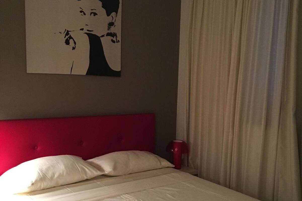 Una habitación de hotel con una cama grande, un cabecero rojo, una colcha blanca, una cortina color beige y un cuadro en la pared.