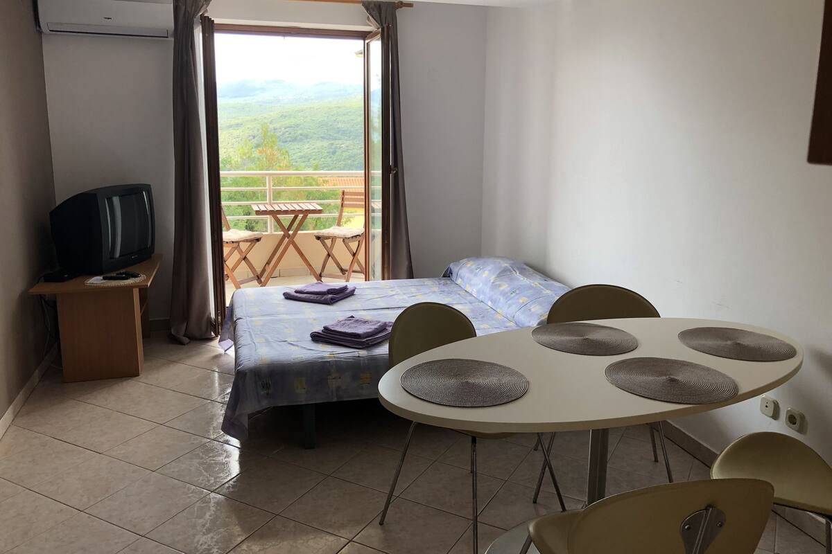 Habitación de hotel con cama, televisión, balcón con mesa y sillas, y vista al exterior.