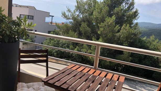 Un balcon avec une table en bois et un siège rembourré, donnant sur une zone boisée et des bâtiments.
