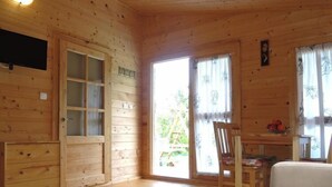 A wooden cabin interior with a wooden door, a television mounted on the wall, a dining table with chairs, and a sofa.