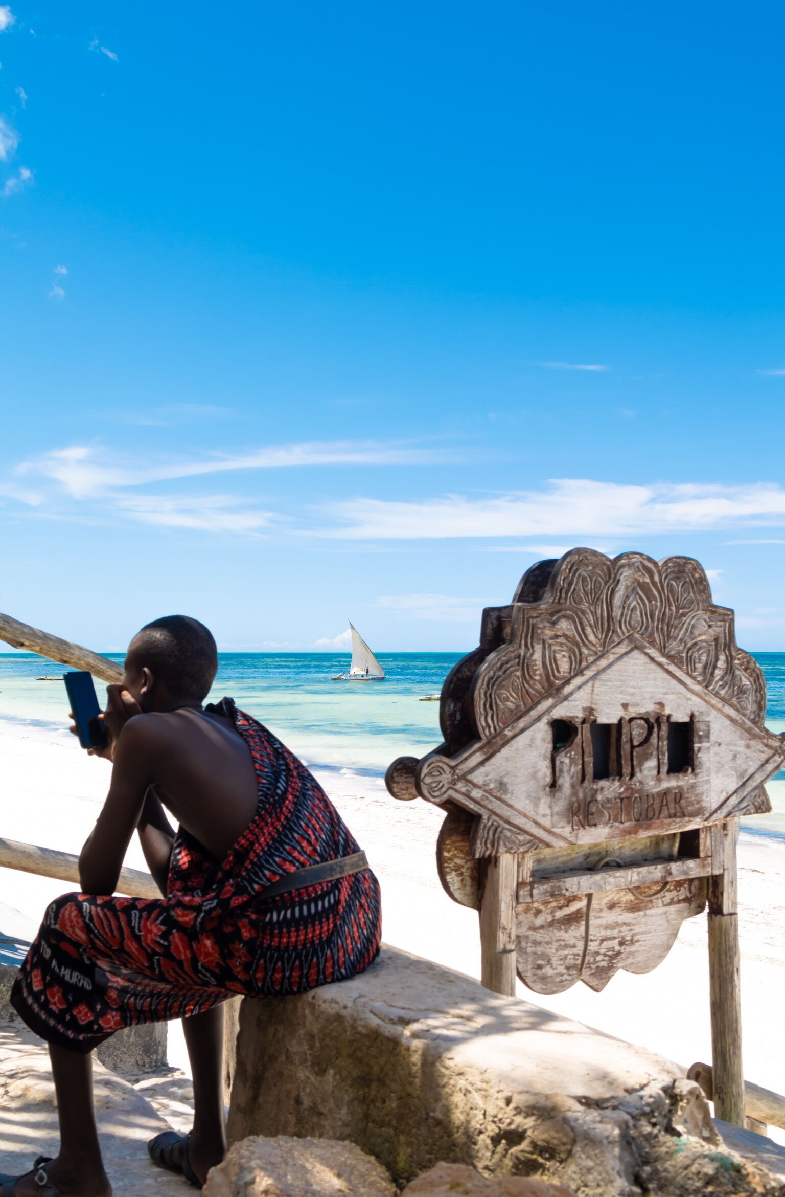 A person sitting on a rock by the beach, a sailboat in the distance, and a wooden sign with the word "RESTOBAR" on it.