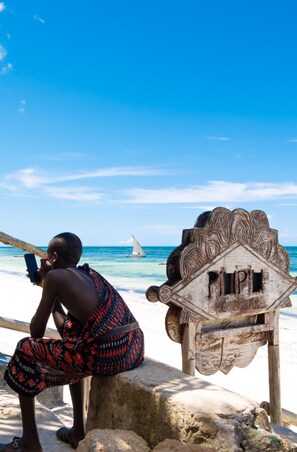 A person sitting on a rock by the beach, a sailboat in the distance, and a wooden sign with the word "RESTOBAR" on it. - Pili Pili House (Jambiani)