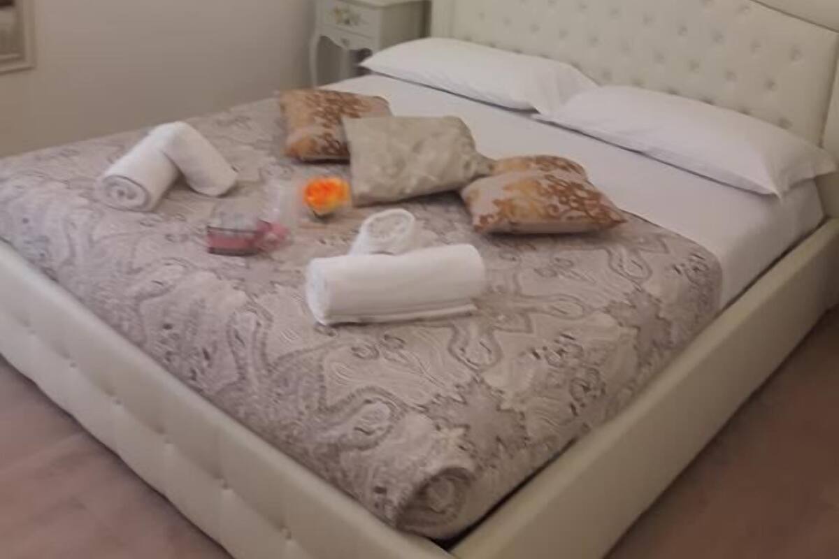 Un lit bien fait, avec des serviettes et des oreillers soigneusement disposés, une table de chevet avec une lampe et un tableau représentant un coucher de soleil accroché au mur.