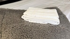 A neatly made bed with a grey textured blanket and a folded white towel on top. - Volontärgatan - Visby Lägenhetshotell (Visby)