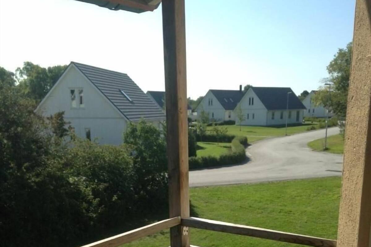 Ein Balkon mit Holzgeländer, einem einzelnen schwarzen Stuhl und Blick auf eine Wohnstraße mit Häusern und Grünflächen.