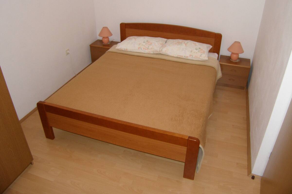 Una cama pequeña y ordenada con estructura de madera y colchón color crema, flanqueada por dos mesitas de noche con lámparas, en una habitación con paredes blancas lisas y suelo de madera.