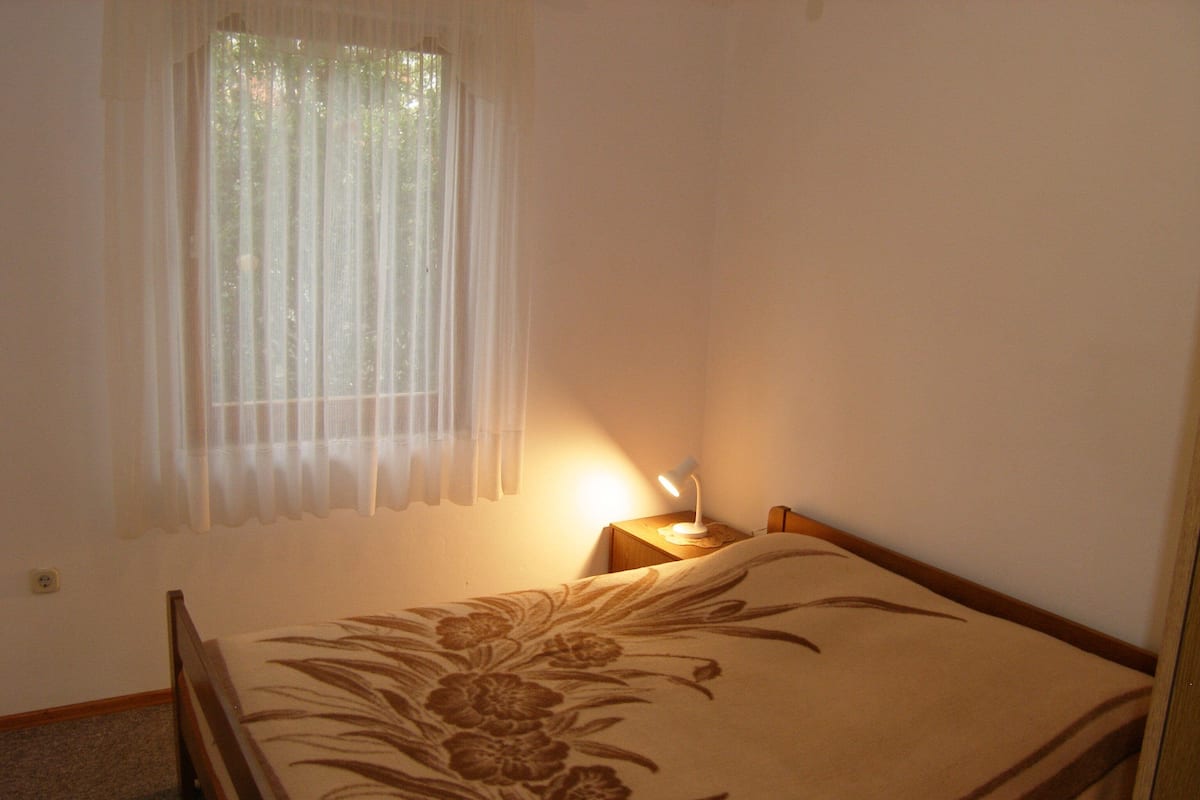 Un dormitorio con cama, una mesita de noche con lámpara, una ventana con cortinas traslúcidas y suelo alfombrado.