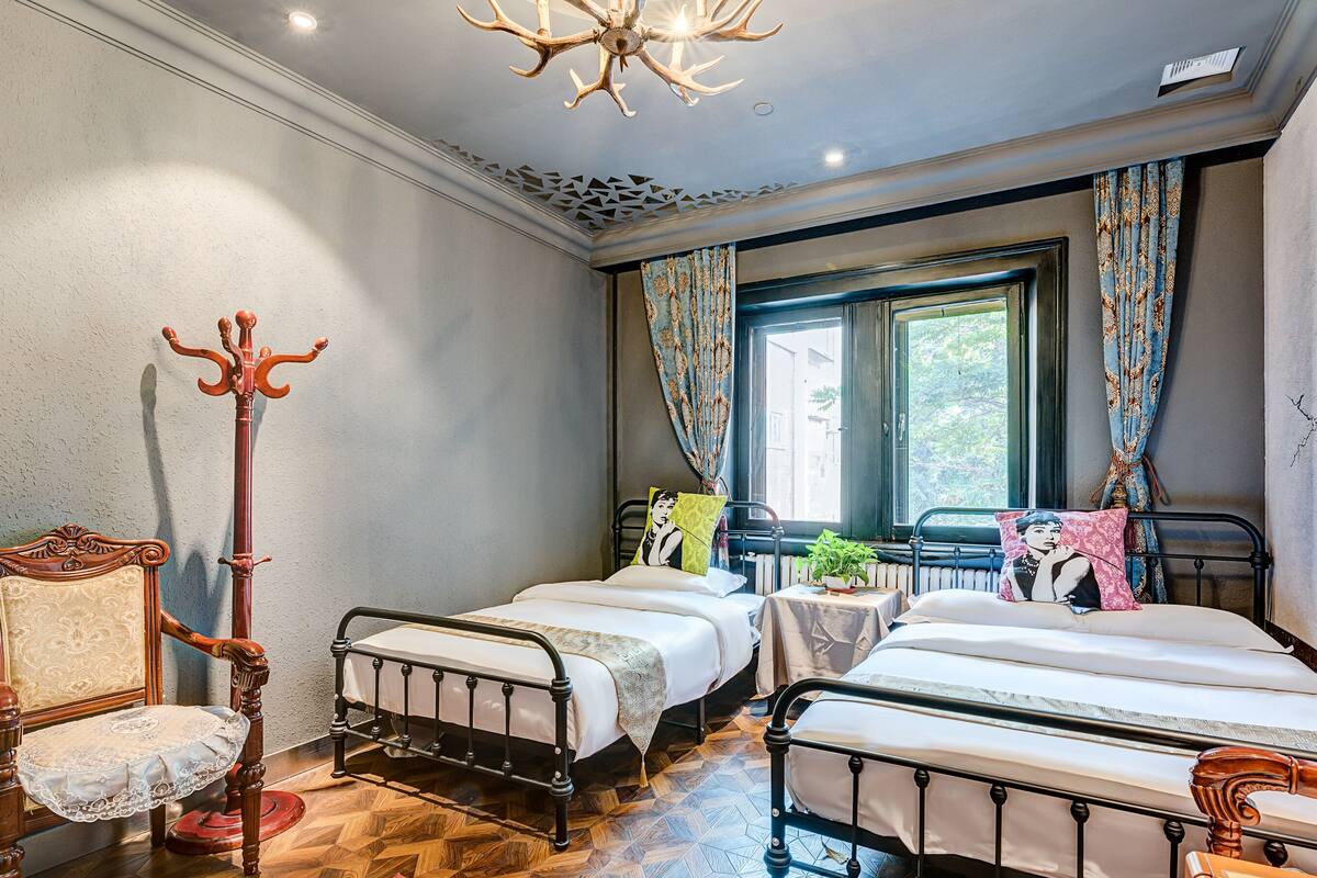 Un dormitorio con dos camas, una silla de madera, un radiador y una ventana con cortinas.