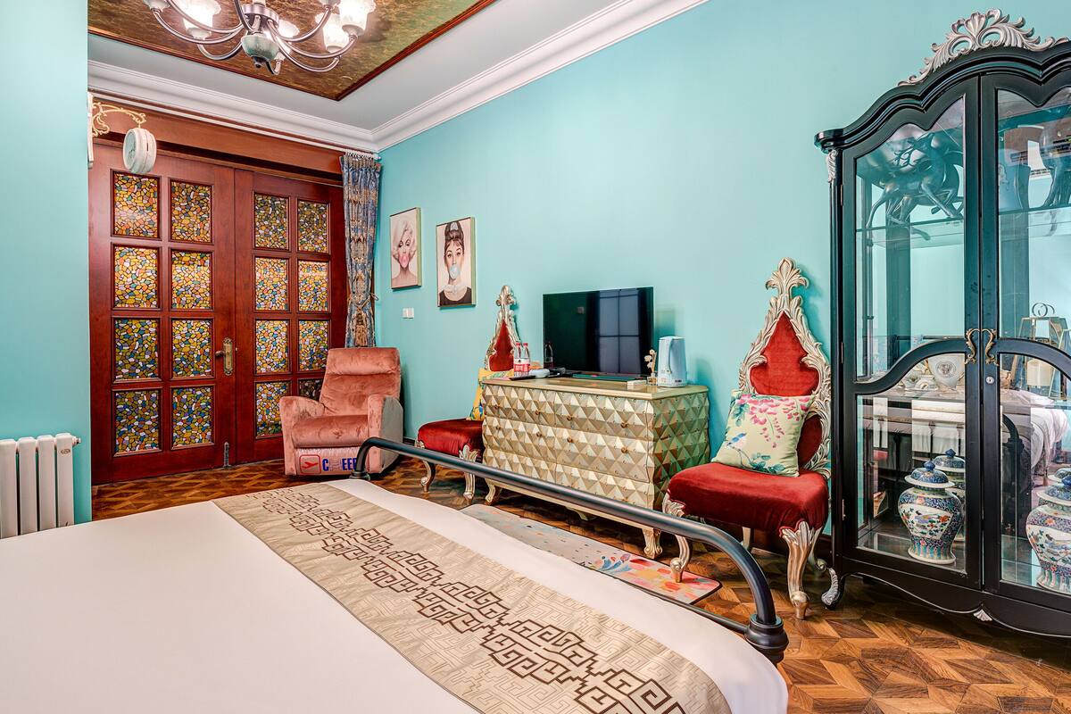 Una habitación con cama, televisión, aparador con puertas de cristal y una silla roja.