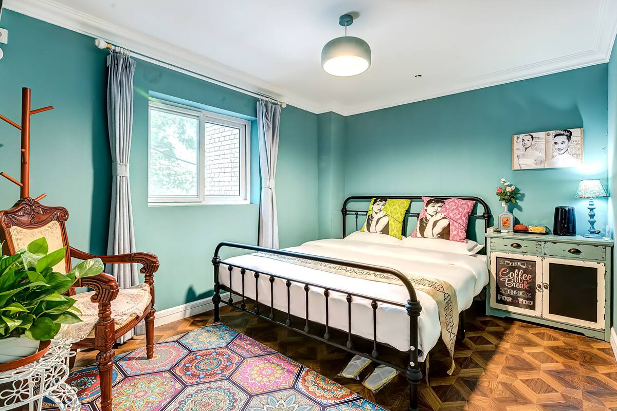 Un dormitorio con una cama, una silla, un mueble bajo, una ventana y una planta.