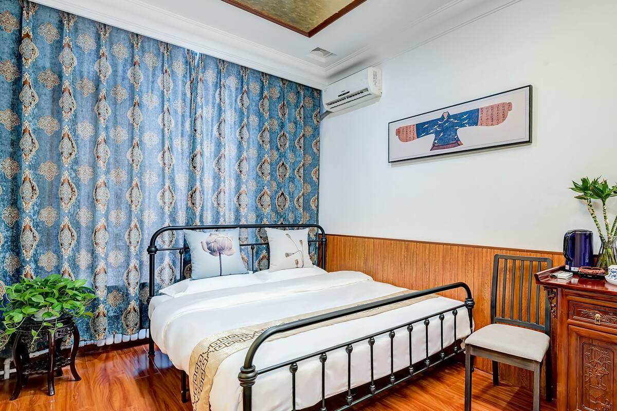 Un dormitorio con una cama, un aparador de madera, una silla, una planta en maceta y un aire acondicionado de pared.