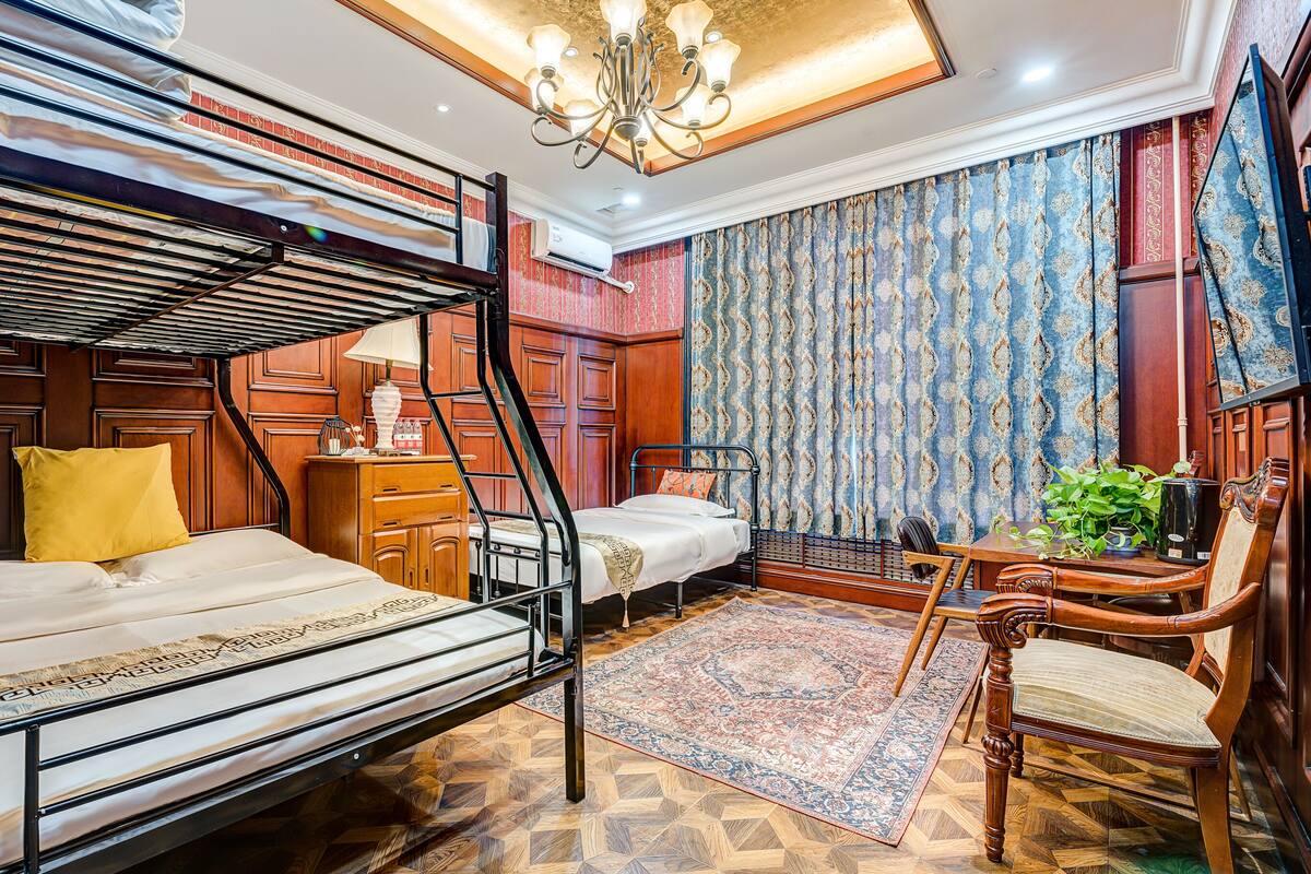 Habitación de hotel con literas, un escritorio con silla, una mesita con planta y un ventanal grande con cortinas.
