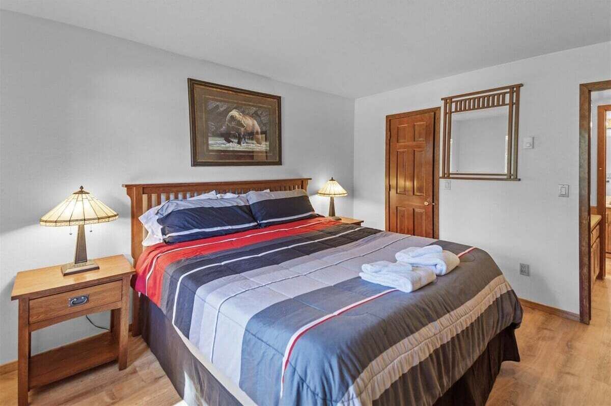 A neatly made bed with striped bedding, two bedside lamps, a wooden nightstand, a framed picture on the wall, and a door with a mirror.