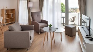 A modern living room with a sofa, armchair, ottoman, and a small table with a plant. There is a TV on a stand, a lamp, and a bookshelf. - DOMITYS - La Majolique (Béziers)