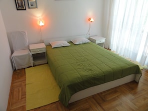 A neatly arranged bedroom with a green bedspread, two bedside lamps, a nightstand, a chair, and a window with curtains. - Cozy apartment Buterer (Rovinj)