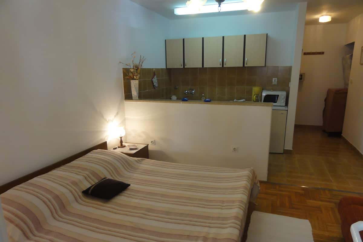 A compact hotel room with a bed, bedside table, lamp, and a kitchenette area with cabinets and a microwave.
