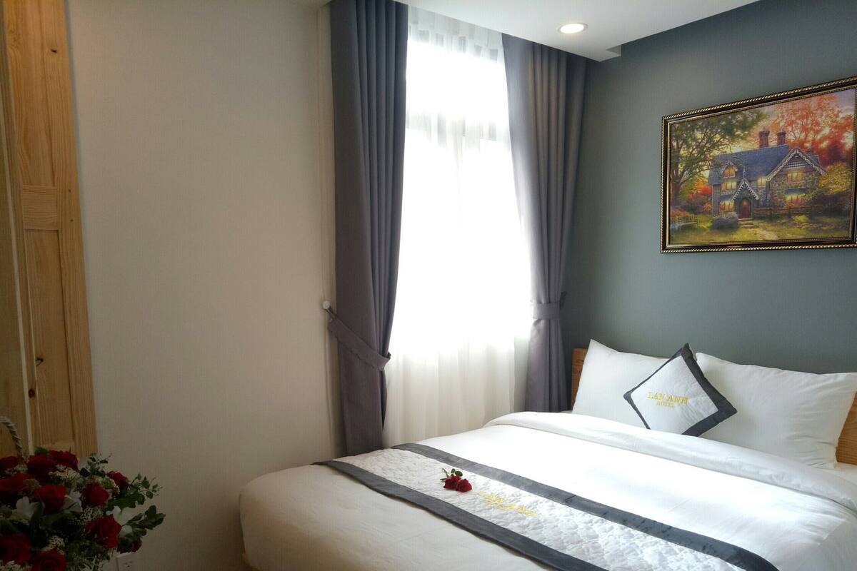 A neatly made bed with a white and gray bedspread, a single pillow with a logo, and a framed painting of a house on the wall.